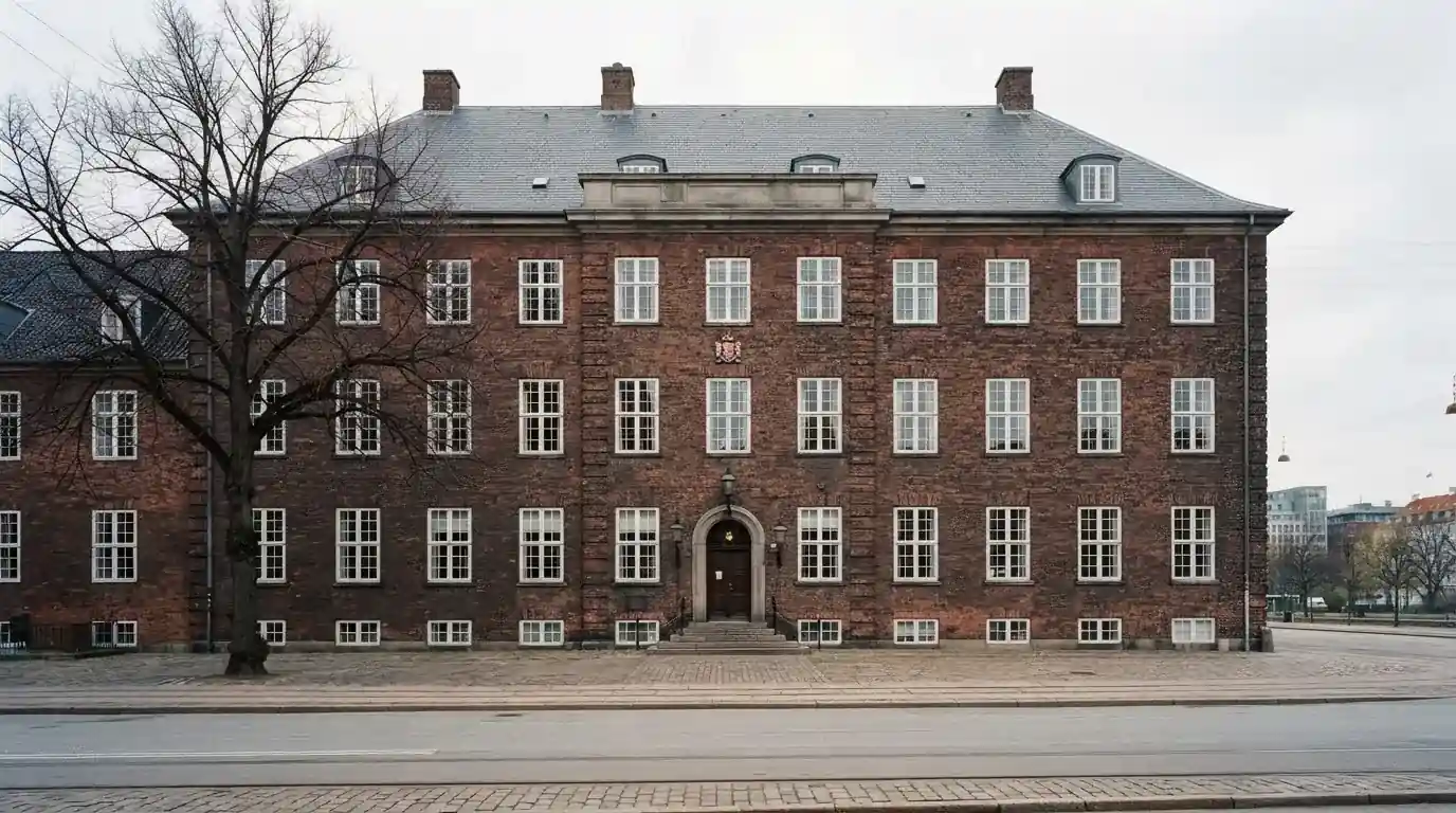 Facade af en gammel administrationsbygning i røde mursten med høje vinduer under skyet himmel, uden personer.